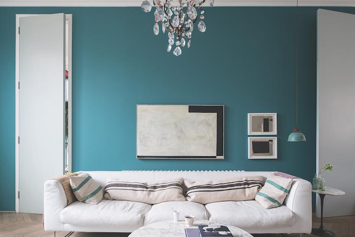 model dnevne sobe v bencinsko modri barvi, steno pobarvano v pastelno modri barvi, beli kavč, okrašen z zelenimi in rjavimi črtastimi bežimi blazinami, marmorna mizica, eleganten lestenec