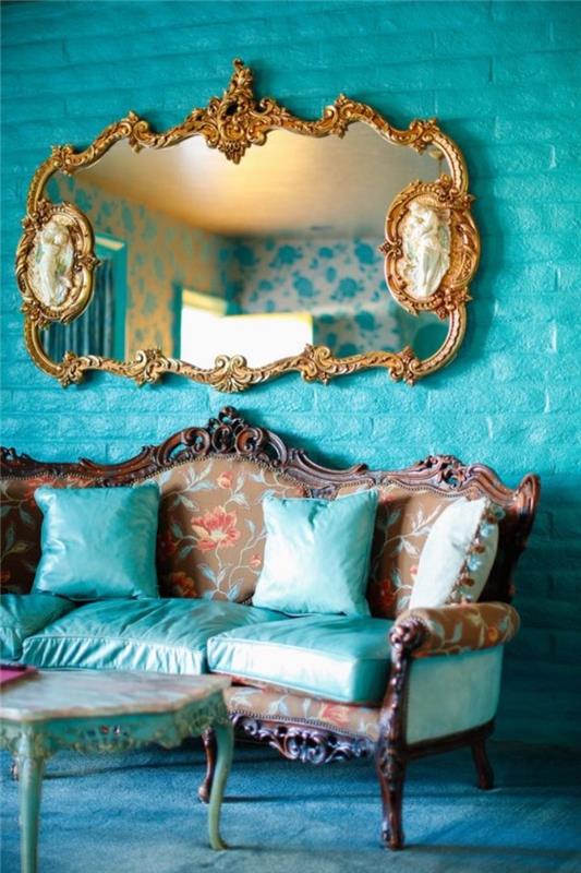 tamsiai žalios spalvos tapybos sienų dekoravimo idėja ir barokinė sofa su dekoratyvinėmis pagalvėlėmis ir pastelinė žalia sėdynė, senovinis kavos staliukas, mėlynas kilimas ir barokinis veidrodis