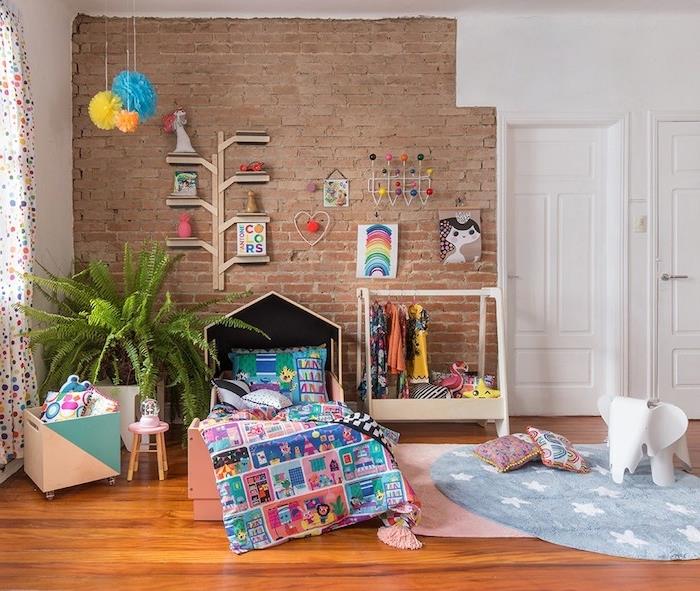 dekoratyvinė plytų siena su medinėmis parketo grindimis, rožinis kilimas ir apvalus kilimas, žaislų skrynia su ratukais, atvira vaikų drabužių spinta