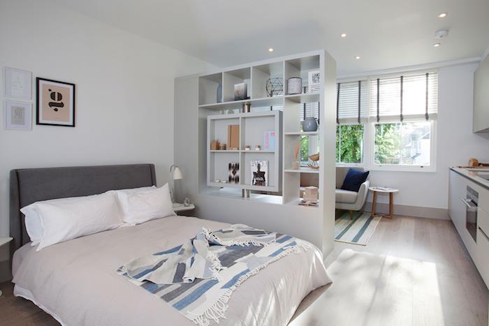 postavitev majhnega stanovanja s sivo -belo posteljo, ločeno od majhnega dnevnega prostora s sivim kavčem, bela kuhinja s črno delovno ploščo