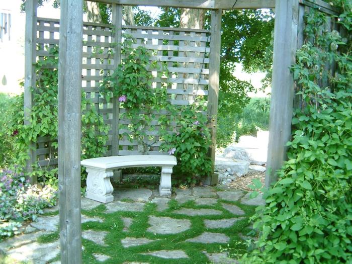 vertikalus sodas ant pilkšvos medinės sienos, sodo paviljono pavėsinė, žemė padengta dideliais pilkais akmenimis, apsupta žalios žolės, išorinė žalia siena, žalia siena