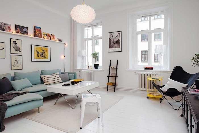 mėlyna kampinė sofa ir baltas sulankstomas stalas šalia, baltos sienos ir dizainerio pakabinama šviesa, ekologiška rėmų siena ir šviesi girlianda skandinaviškam „cocooning deco“