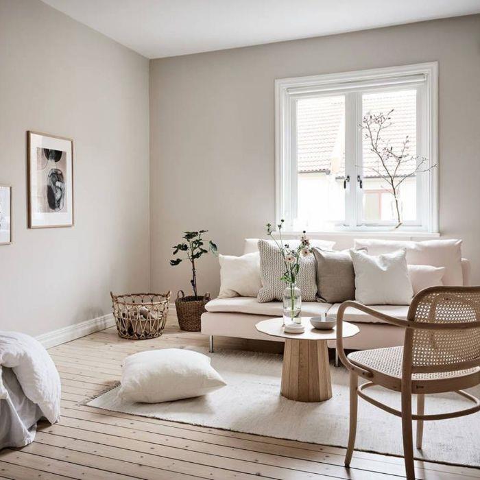 perlamutrinės pilkos sienos smėlio spalvos sofa, dekoruota pagalvėlėmis baltas kilimas minimalistinis kavos staliukas, kėdė originali šviesaus medžio parketas