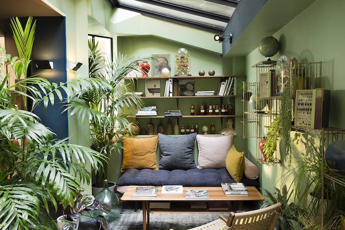zelena barvna stena, lesena miza in sivo -zelena zofa, siva, bela in rumena blazina, množica rastlin v lončkih na tleh, tropski dekor