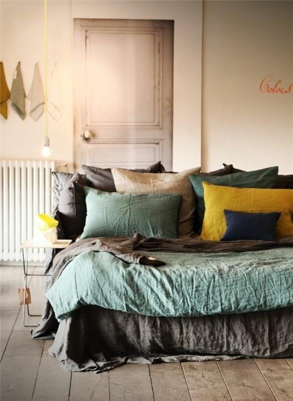 šviesiai žalios ir rudos, tamsiai mėlynos geltonos spalvos pagalvės ant lovos su atitinkamais užvalkalais, kambaryje su šviesiai rudomis medinėmis grindimis, akcento spalva su pilkomis sienomis