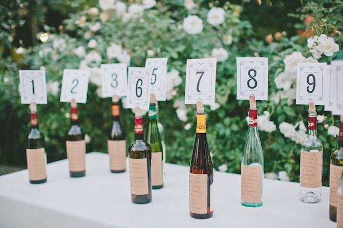 üzerlerine konuk etiketleri yapıştırılmış kişiselleştirilmiş şarap şişeleri ve bir mantar yuvasında masa numarası, diy düğün masası planı