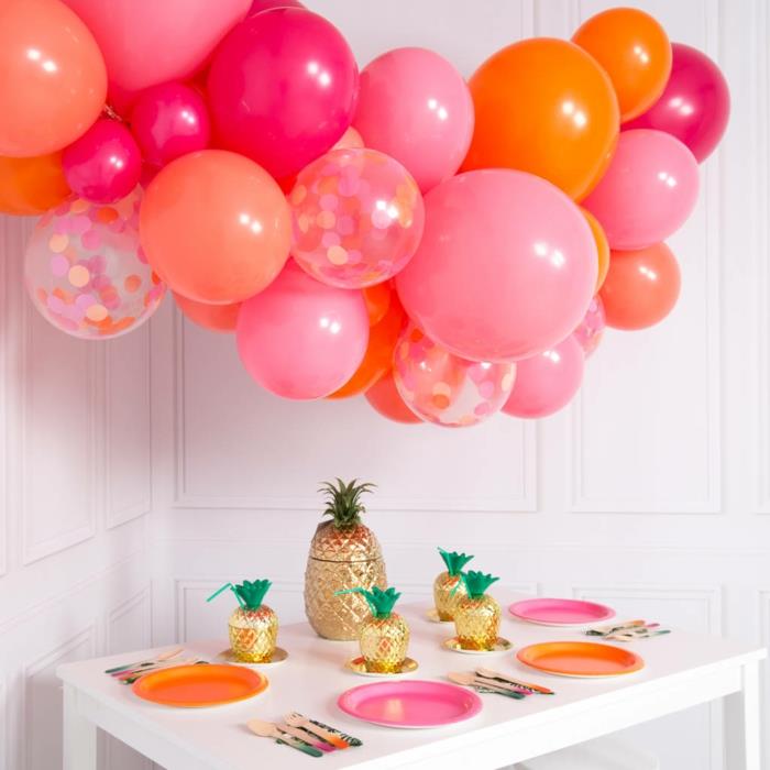 rožinės ir oranžinės spalvos balionų debesis, auksiniai ananasai, balionų spalvų lėkštės, mažas baltas stalas