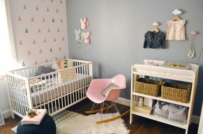 kūdikio darželis su mėlynai pilkais dažais vienoje sienoje, o šviesiais, raštuotais tapetais kitoje, rožinė supamoji kėdė, įvairios rožinės ir pilkos spalvos dekoracijos