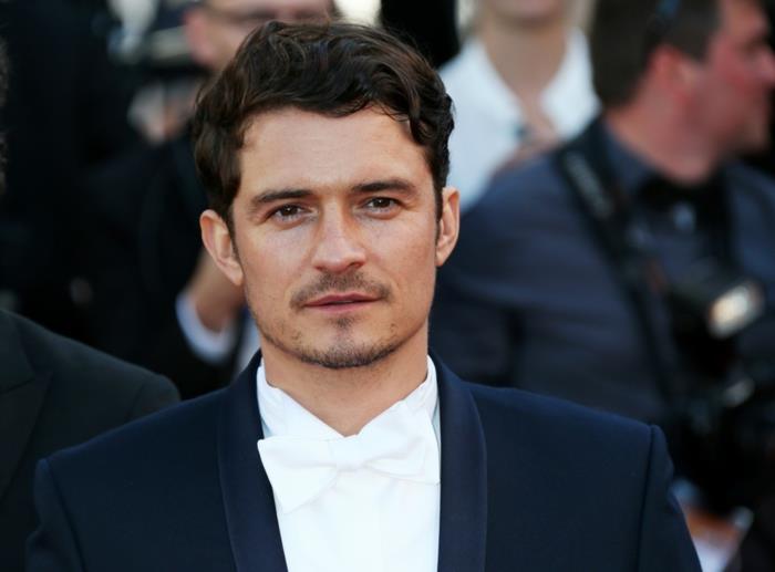 kratki kodrasti lasje, ki jih nosi Orlando Bloom, v beli svečani majici, ki se ujema z metuljčkom in črnim smokingom, kratko brado in brki