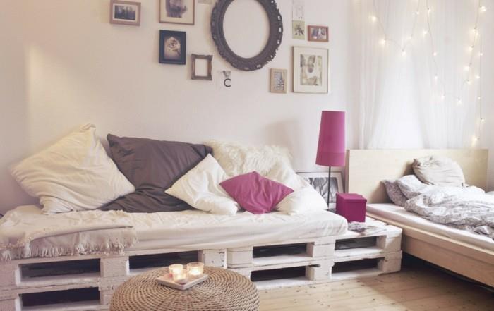 pasakų žiburiai ir paveikslų rėmai, dekoruojantys kambarį su lova ir baltomis užuolaidomis, kurioje yra didelė sofa, padaryta iš padėklų ir padengta pagalvėlėmis