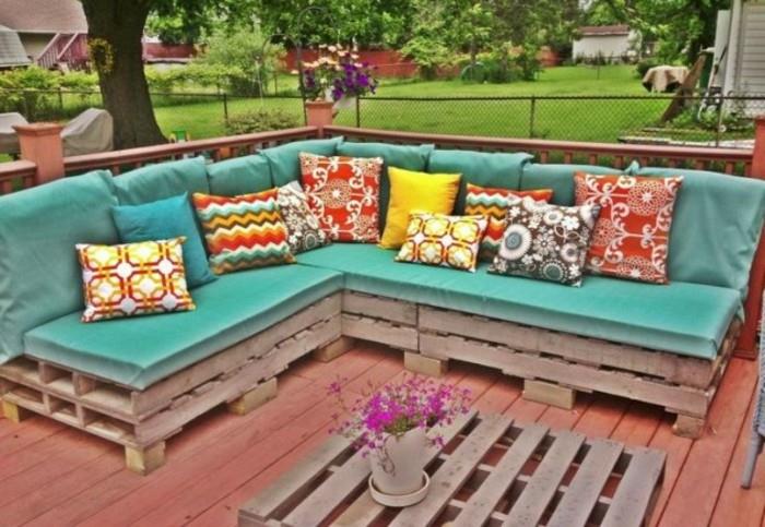 kampinė sofa pagaminta iš padėklų, padengta turkio spalvos putų čiužiniais ir papuošta įvairių dydžių įvairiaspalvėmis raštuotomis pagalvėmis.