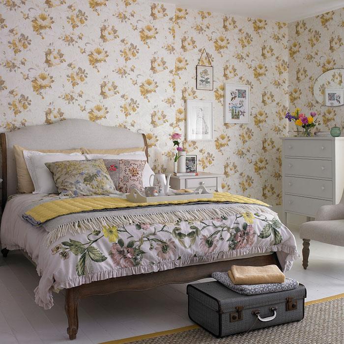 çiçek desenli duvar kağıdı modeli, beyaz zemin üzerine sarı çiçekler, ahşap yatak, hafif parke, yatak ucu olarak gri vintage sandık, minderler, çiçek desenli battaniye