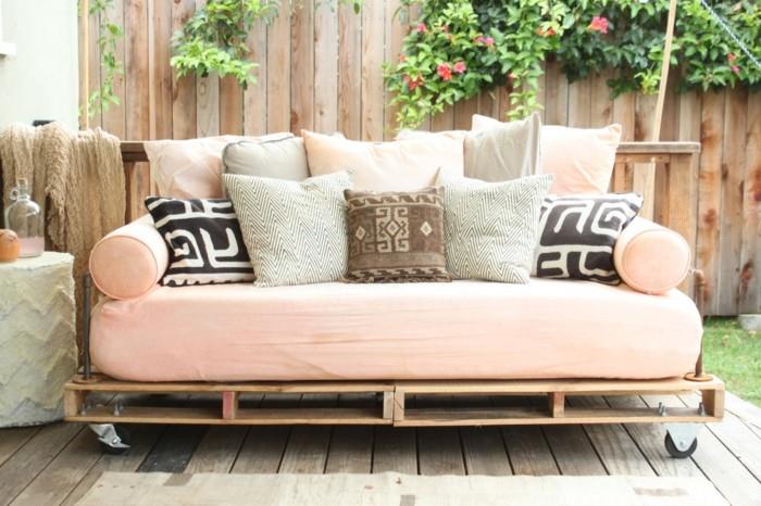 šviesiai rožinis čiužinys, dengiantis sofą iš medinių padėklų, dekoruotas daugybe pagalvėlių, rožinės ir pilkos, juodos ir baltos, rudos ir smėlio spalvos
