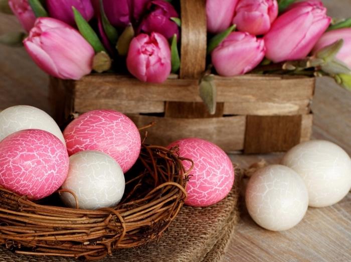 medinis stalas su krepšeliu su rožinėmis tulpėmis ir spalvingų rožinės ir baltos spalvos kiaušinių lankas su sulaužytu kiaušinių dizainu