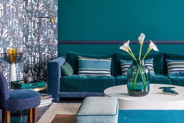 benzininis mėlynas svetainės modelis, anties spalvos sienų dažai, mėlynos, žalios ir baltos pagalvėlės, tamsiai mėlyna ir povo mėlyna sofa, atogrąžų rašto tapetai, minimalistinis baltas kavos staliukas