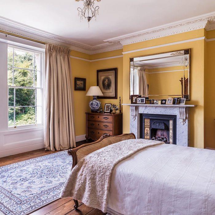 rumena barva spalnice za odrasle v starinski sobi, lesena postelja in starinska komoda, belo posteljnina, rjavi parket, dizajnerski kamin