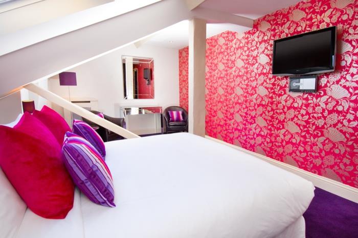 moderna spalnica, dekor za spalnico, ideja za okrasitev spalnice, stenska tapeta s satenskim zaključkom v rdeči in sivi barvi z arabesknimi vzorci, soba pod podstrešjem s poševnimi stropi, preproga v vijolični barvi, posteljne blazine v fuksiji in vijolični barvi