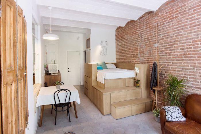 lova ant medinės platformos šalia plytų sienos, valgomojo zona ir dizainerio odinė sofa, 25 m2 studijos išplanavimas