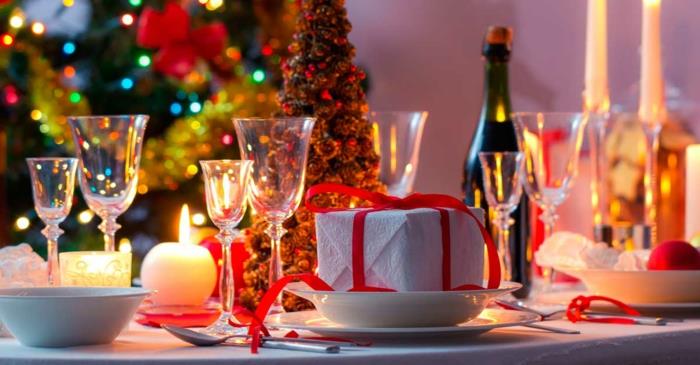 vyno taurės, dovana, įvyniota į baltą popierių, perrišta raudona juostelė, Kalėdų eglutė kaip centras, didelė Kalėdų eglutė su šviesiomis girliandomis