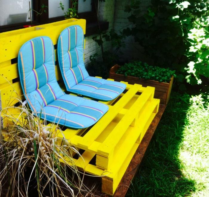 padėklo suoliukas, nudažytas ryškiai geltona spalva ir padengtas dviem pastelinės mėlynos ir geltonos spalvos čiužiniais, paletiniai sodo baldai, pastatyti medžio pavėsyje
