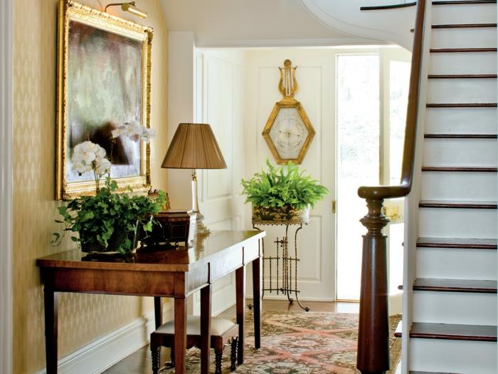 medinis stalas prieškambariui, lempa su šešėliu, aukso rėmo tapyba, senovinis kilimas, žali augalai
