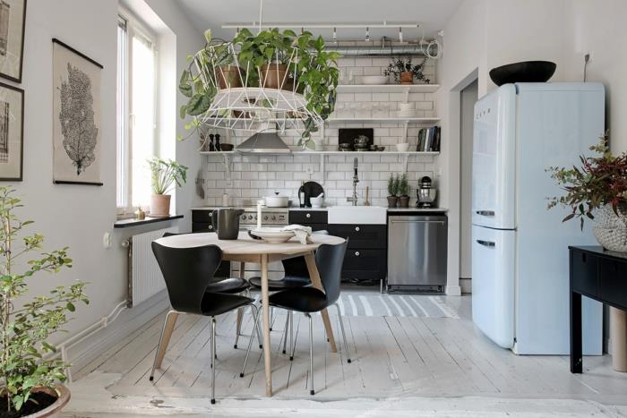 maža balta virtuvė, apvalus stalas su juodomis kėdėmis, balintos parketo grindys, pastelinės mėlynos spalvos šaldytuvas, lentynos, metro plytelės, keli augalai