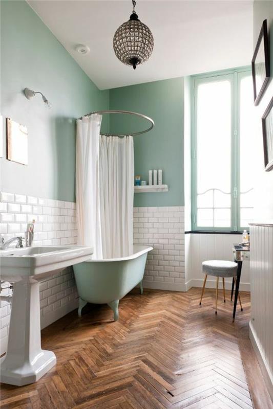 mažas vonios kambarys su pistacijų žalia vonia ir baltomis užuolaidomis ant apvalaus metalo virš vonios