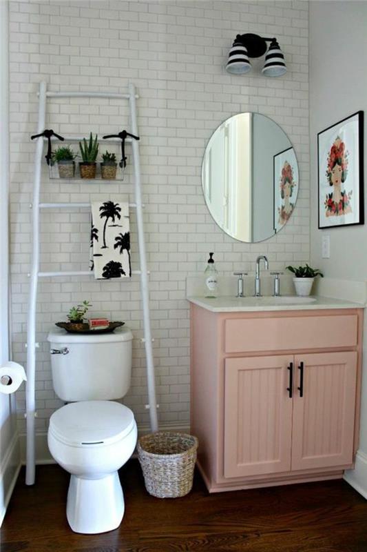 majhna kopalnica iz bele opeke z rožnato omarico z žensko v barvah in okroglim ogledalom brez okvirja
