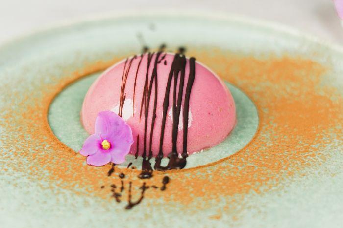 panakotos nuotrauka rožinėje desertinėje lėkštėje, šokolado puošmena ir valgomoji gėlė marmurinėje lėkštėje