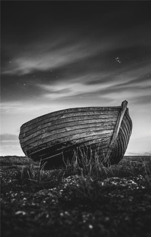 poetična črno -bela podoba osamljenega čolna, opuščenega na obali pod plesnimi oblaki