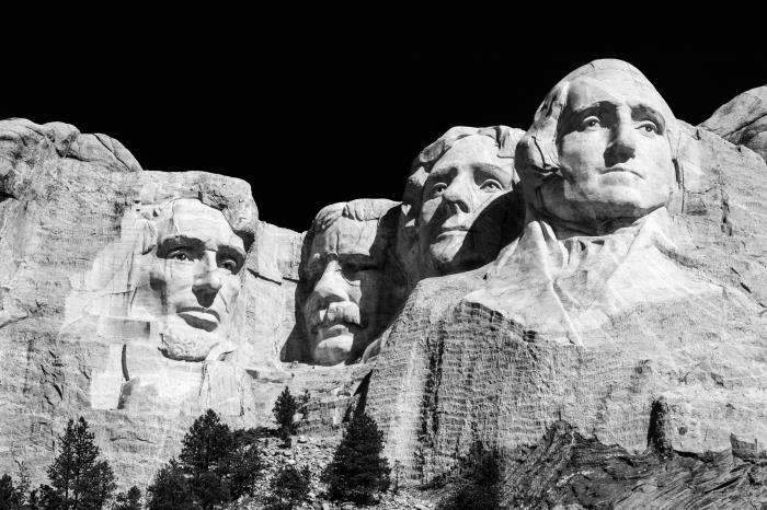 črno -bela podoba gore Rushmore s štirimi predsedniki, vklesanimi v skalo, obrisani na črnem ozadju neba