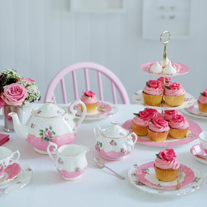 gražus pyragas iš mažų pyragų, rožinės ir baltos spalvos indai, maža rožių puokštė, baltas stalas, rožinė kėdė