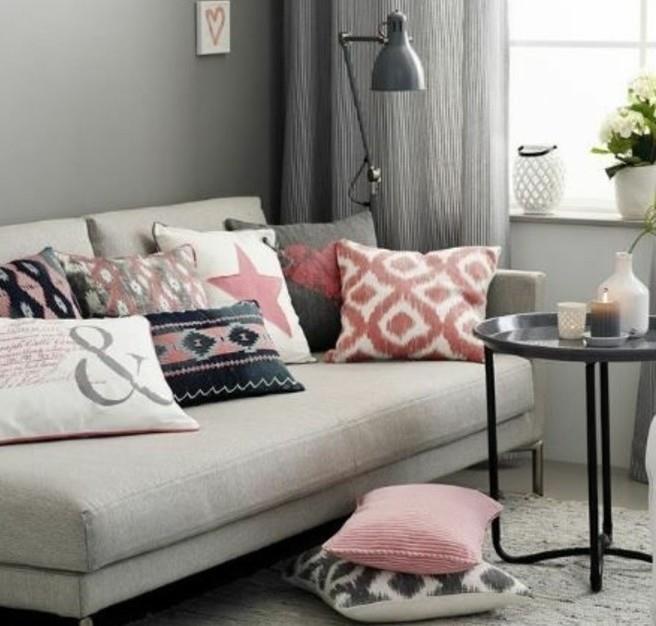 įvairios rožinės, baltos ir pilkos spalvos pagalvėlės ant šviesiai pilkos sofos, šalia juodo metalo kavos staliuko, svetainės dažų spalvos, pilkos sienos ir užuolaidos