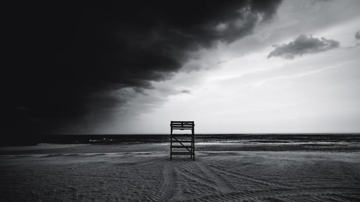 črno -bela morska pokrajina zapuščene plaže z osamljeno postajo reševalcev in viharnim nebom, ki je na pol pokrito z oblaki