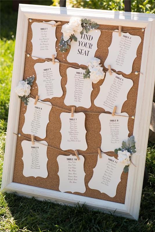 stalo planas vestuvėms baltas rėmas ir kamštinis fonas su baltomis etiketėmis ir svečių vardais juodomis raidėmis, tvirtinami mediniais skalbinių segtukais