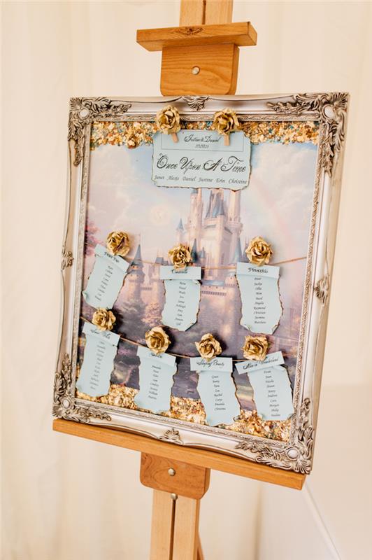 užburtas pilies stalo vestuvių stalo planas baroko rėmelyje, medinis molberto stovas, pastelinės mėlynos etiketės su svečių vardais ir auksinėmis rožėmis