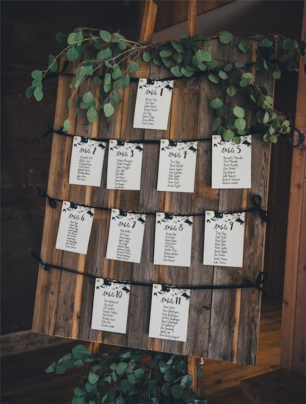vestuvių stalo planas rudomis vintažinėmis medinėmis lentomis ir mažais baltais svečių sąrašais, kaligrafija juodomis raidėmis, žalia girlianda