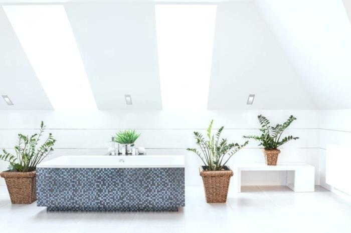 balta stačiakampė vonia, padengta mėlynos ir pilkos spalvos mozaikinėmis plytelėmis, lubos su dviem stačiakampiais langais, šviesi erdvė, vonios kambarys, 4 dideli žalių augalų vazonai
