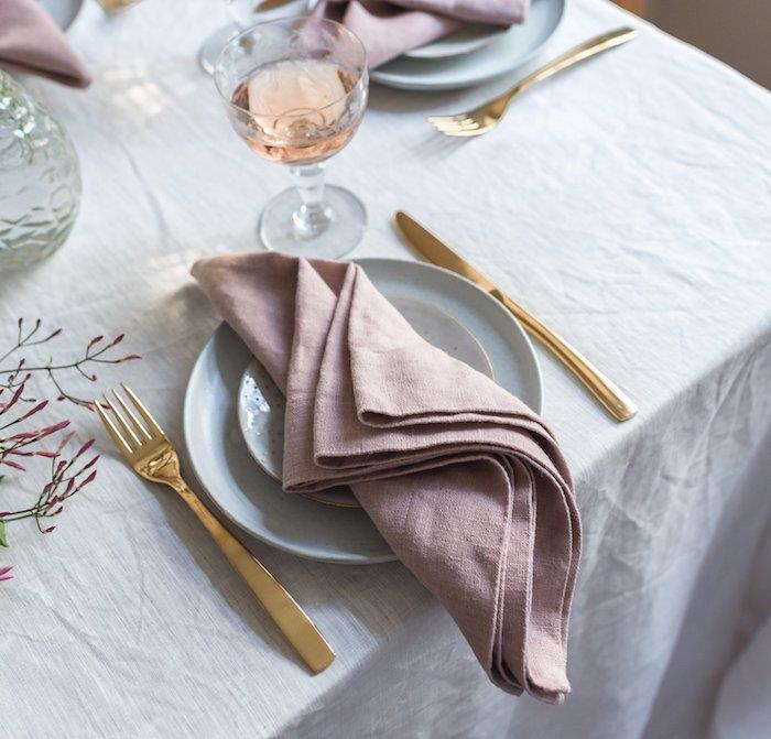 lengvai sulankstoma šviesiai rožinė servetėlė ant baltos staltiesės, auksiniai stalo įrankiai, lengva servetėlės ​​sulankstymo idėja