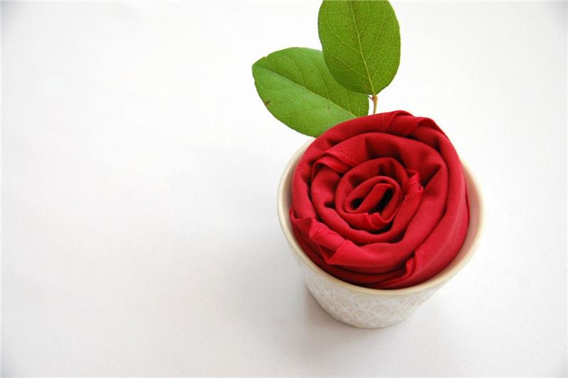 pavyzdys, kaip sulankstyti audinio servetėlę rožės pavidalu stikle su natūraliais žaliais lapais, romantiška stalo puošmena
