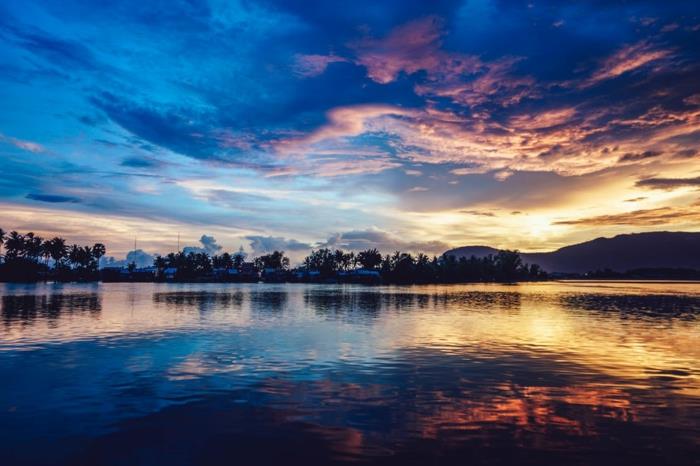rojaus kraštovaizdis mėlynos ir violetinės spalvos, šiek tiek banguoti vandenys, horizonte esantys palmių siluetai, stebuklingi atspalviai, purpuriniai kalnai