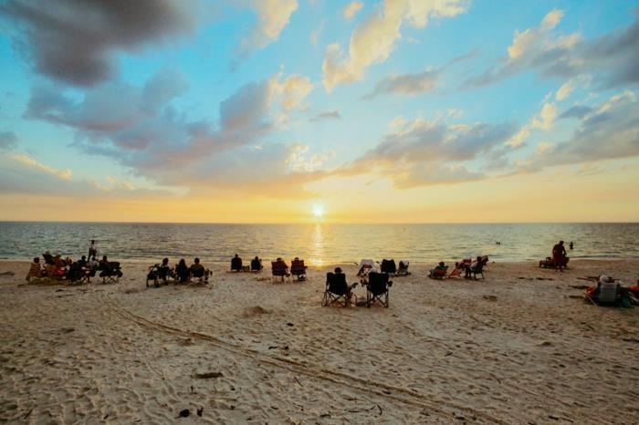 dangiškoji paskirties vieta, paplūdimys saulėlydžio metu, mėlynas dangus, ant gultų gulintys turistai, pėdsakai ant smėlio