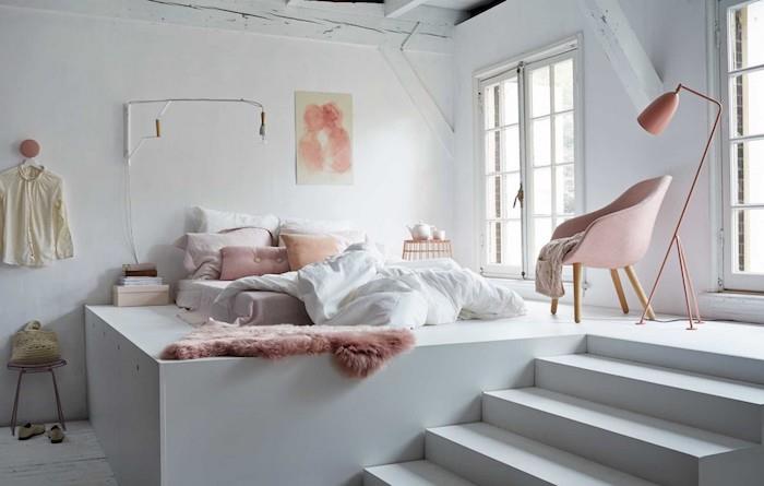 „Cocooning“ lovos čiužinys ant baltos platformos, su pagalvėlėmis ir rožiniu pledu, baltos sienos, rausvas fotelis, higge atmosfera, romantiškas studijos miegamojo dekoras
