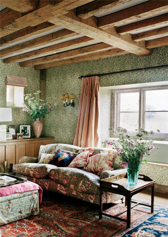 dekor podeželske hiše, cvetlične zofe, otomanska z rustikalnim tapeciranjem, leseni tramovi, roza zavesa, steklena vaza s poljskim cvetjem