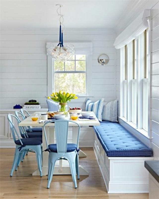 perdažykite virtuvę, kambarį karališkos mėlynos ir baltos spalvos, namo stilių, iš kurio atsiveria vaizdas į jūrą, pastelinės mėlynos spalvos metalines kėdes su karališkos mėlynos spalvos pagalvėlėmis, baltai dažytą medinę sofą su didele stačiakampio formos pagalve karališkos mėlynos spalvos, lygų parketą rudos ir smėlio spalvos, karališkai mėlyną metalą šviestuvas ir gėlių raštai iš skaidraus balto stiklo