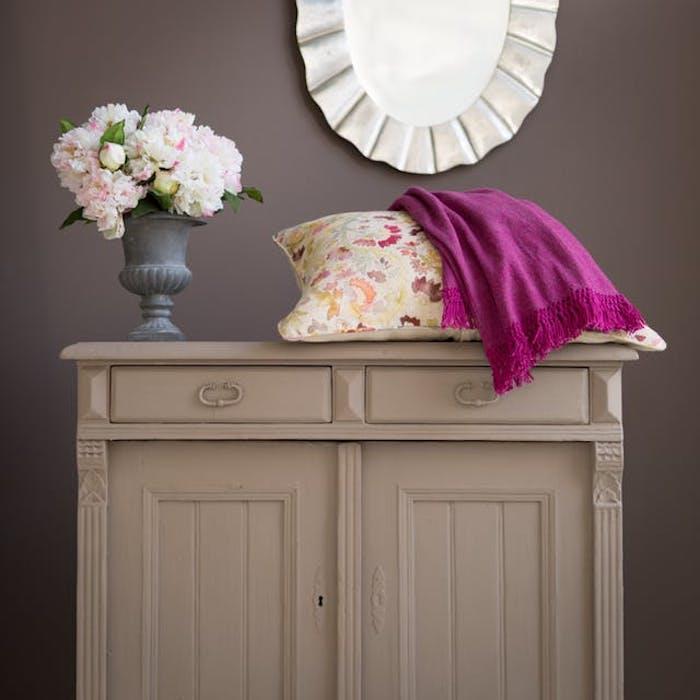 prenovljen kos pohištva, predalnik prebarvan v sivo, vijolično odejo in pisano blazino s cvetličnim potiskom, šopek rož, temno siva stena, okroglo ogledalo