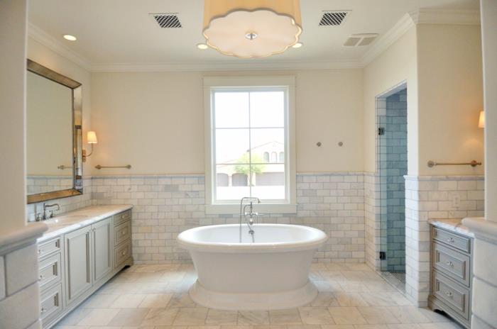 smėlio spalvos lubų šviestuvas, vonia erdvės centre, stačiakampis įrėmintas veidrodis, praustuvai, travertino vonios kambarys