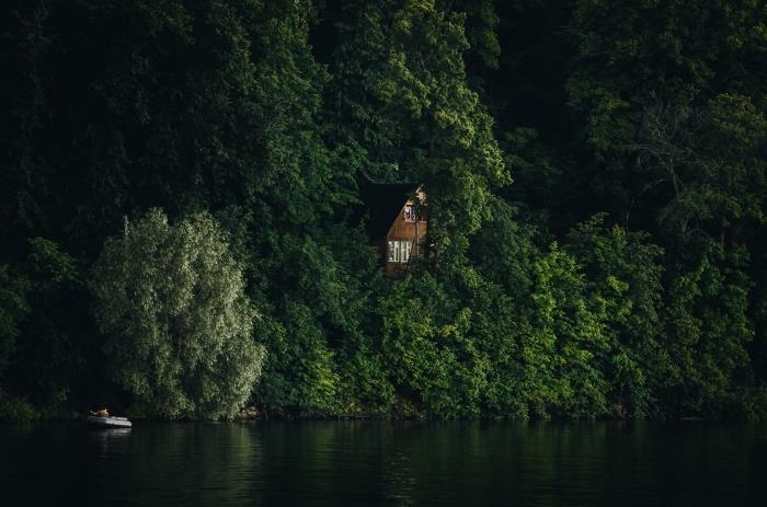 dviejų aukštų medinis namas, pastatytas miške virš ežero, valtis vandenyje atspindinti žalius medžius