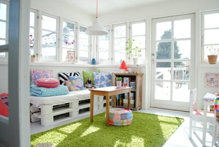 „pasidaryk pats“ sofa, pagaminta iš baltų padėklų ir dekoruota įvairiaspalvėmis pagalvėlėmis, šviesiame kambaryje, su keliais langais ir stiklinėmis durimis