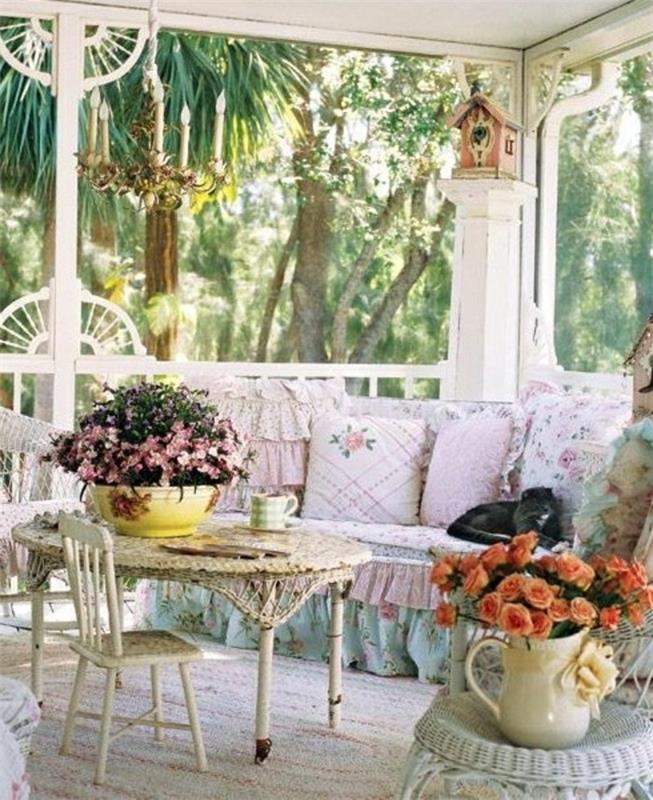 küçük sandalyeli beyaz yuvarlak masa, uçuk mavi ve pembe örtülü kanepenin yanında, uçuk pembe minderlerle süslenmiş, fırfırlar ve çiçek desenli, sundurma dekoru, birçok dekoratif obje